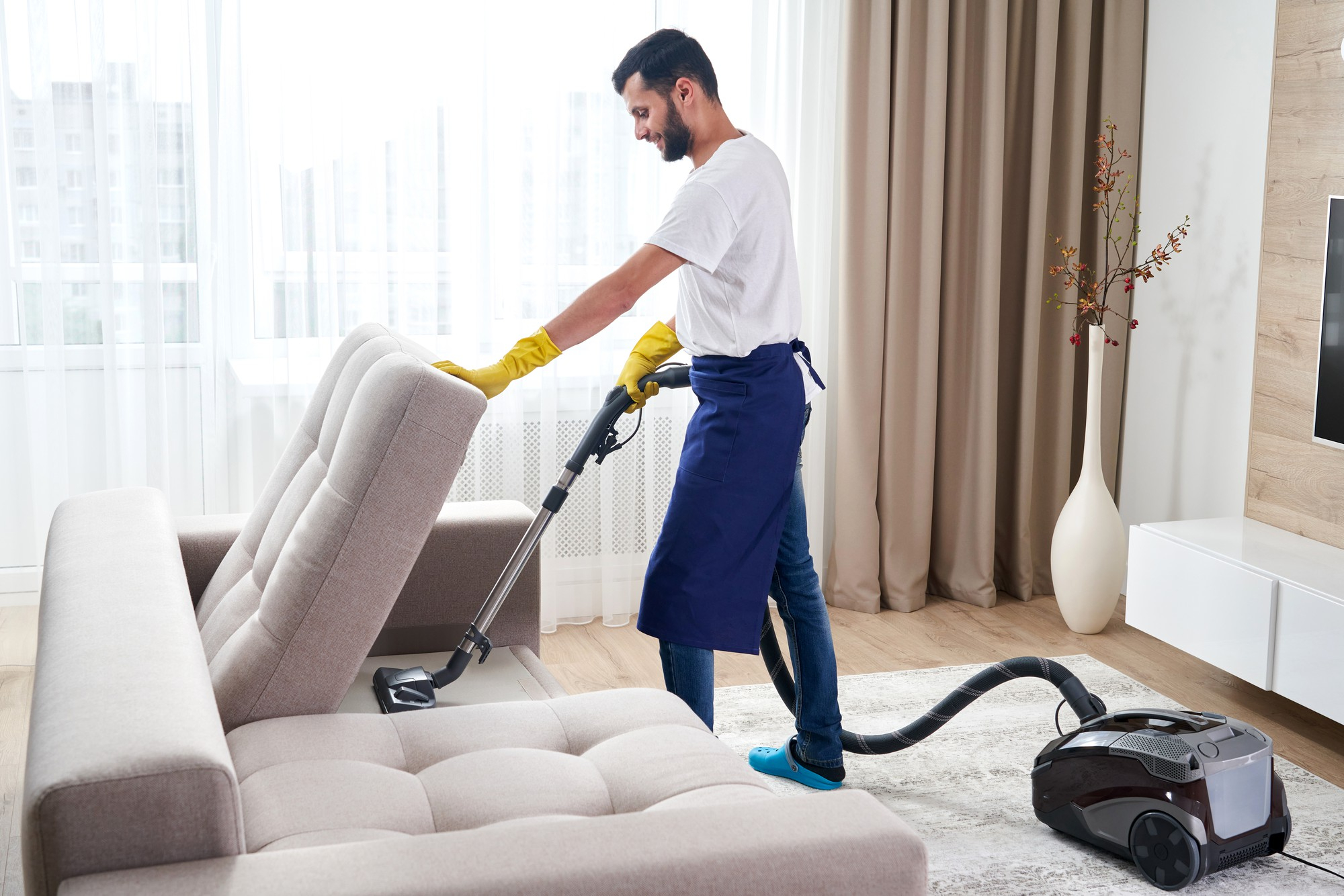 Cleaner Dallas professional performing deep cleaning and vacuuming under a sofa in a modern living room